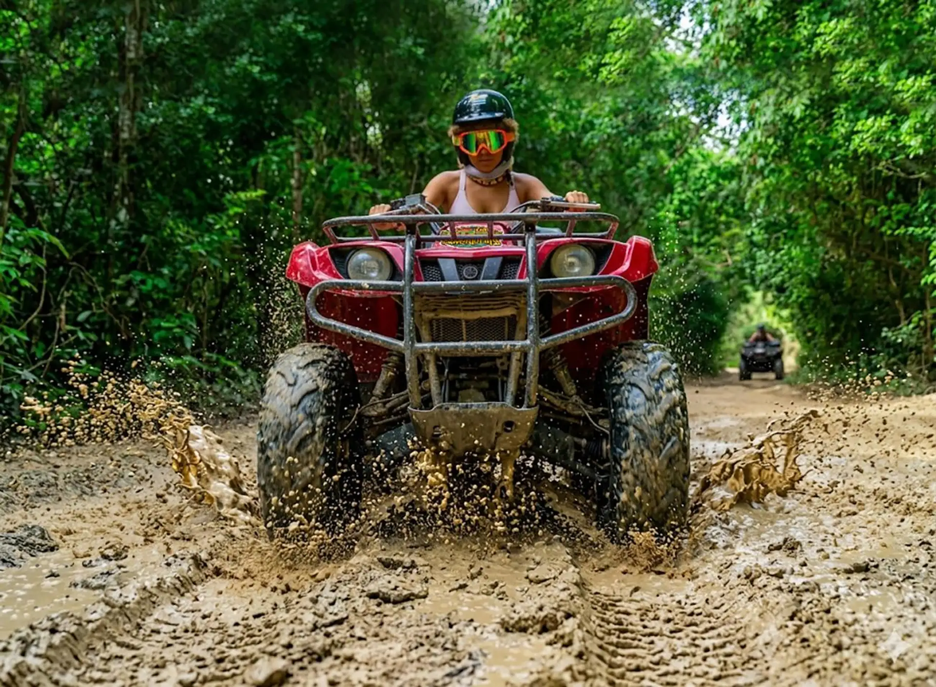 Helmeted rider driving a red ATV through a muddy jungle trail